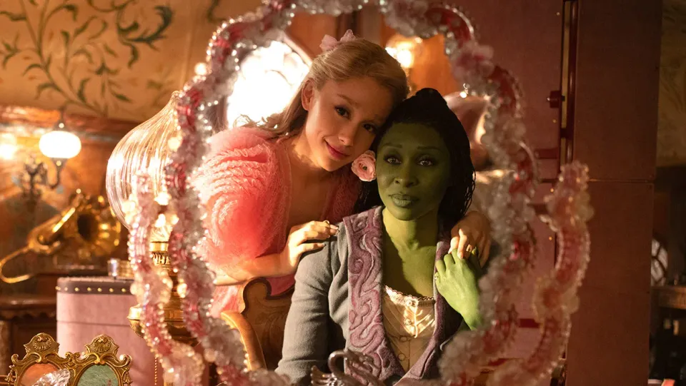 Ariana Grande and Cynthia Erivo star in Wicked. They lean close together to look in a mirror.