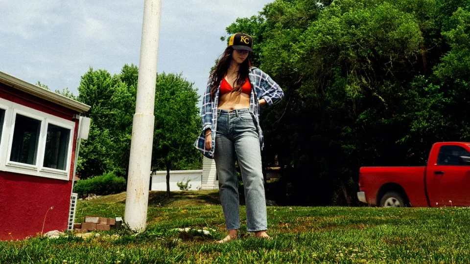 Katie Crutchfield, aka Waxahatchee, stands in the middle of greenery in a shirt over a red bra. She wears a ball cap and 