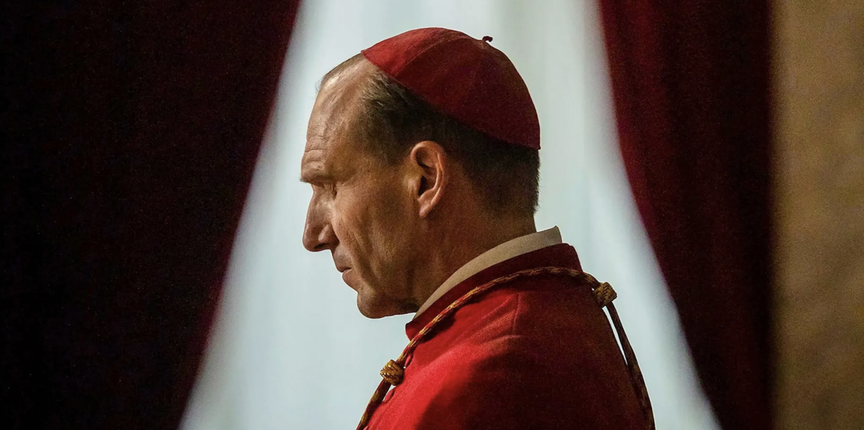 Ralph Fiennes, seen from the side, dresses in full Catholic Cardinal regalia. His eyes are closed in prayer. Or annoyance.