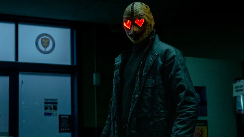 The Heart Eyes Killer stands in a darkened room. Their red heart eyes glow in their otherwise yellow mask.