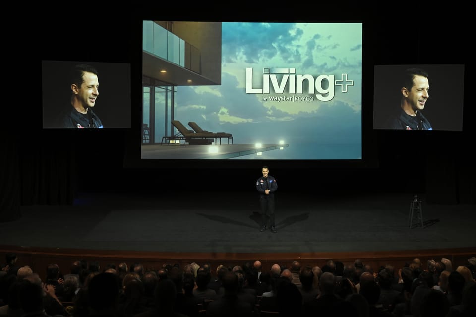 Kendall Roy, tiny, in a flight suit, stands in front of enormous screens showing his face and the logo for Living+.