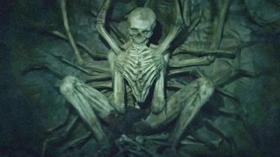 A strange skeleton, its ribcage burst open, tendrils (or arms??) reaching down to it, sits trapped in a cave wall.