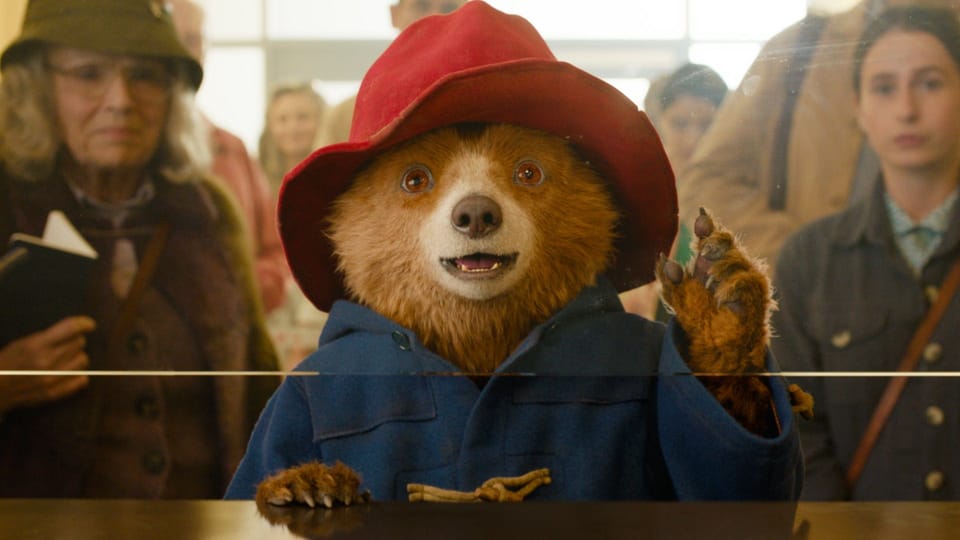 Paddington checks in at customs, the Brown family behind him. He is, as always, a brown bear in a red hat and blue coat.