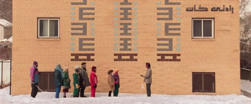 A group of seven people, all in winter wear, listens to a tour guide in red earmuffs in a wide shot in front of a brick build