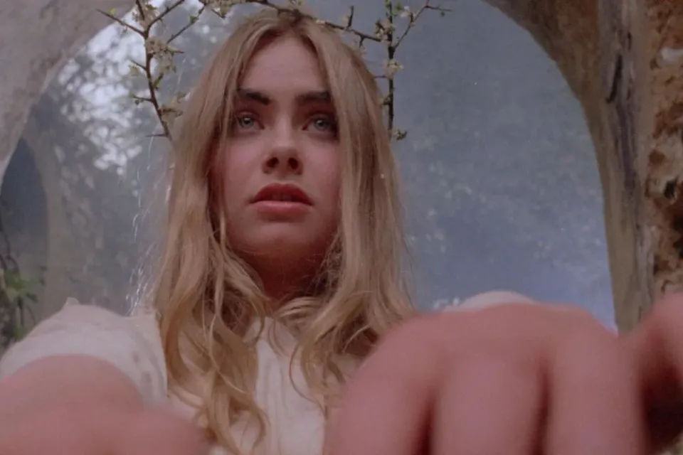 A white, blonde woman with blue eyes, clad all in white, wearing a crown of sticks, leans toward camera in close-up.