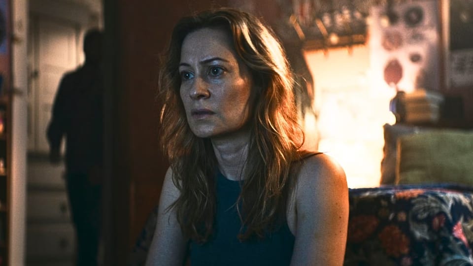 A woman watches something intently on a screen that is unseen to us. She sits on the floor. Behind her, a masculine figure in silhouette stands in the doorway.